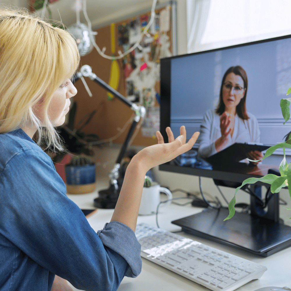 A woman sits at a desk with plants, gesturing while having a video call with another woman who appears on the computer screen, both engaged in a conversation in a home office setting.