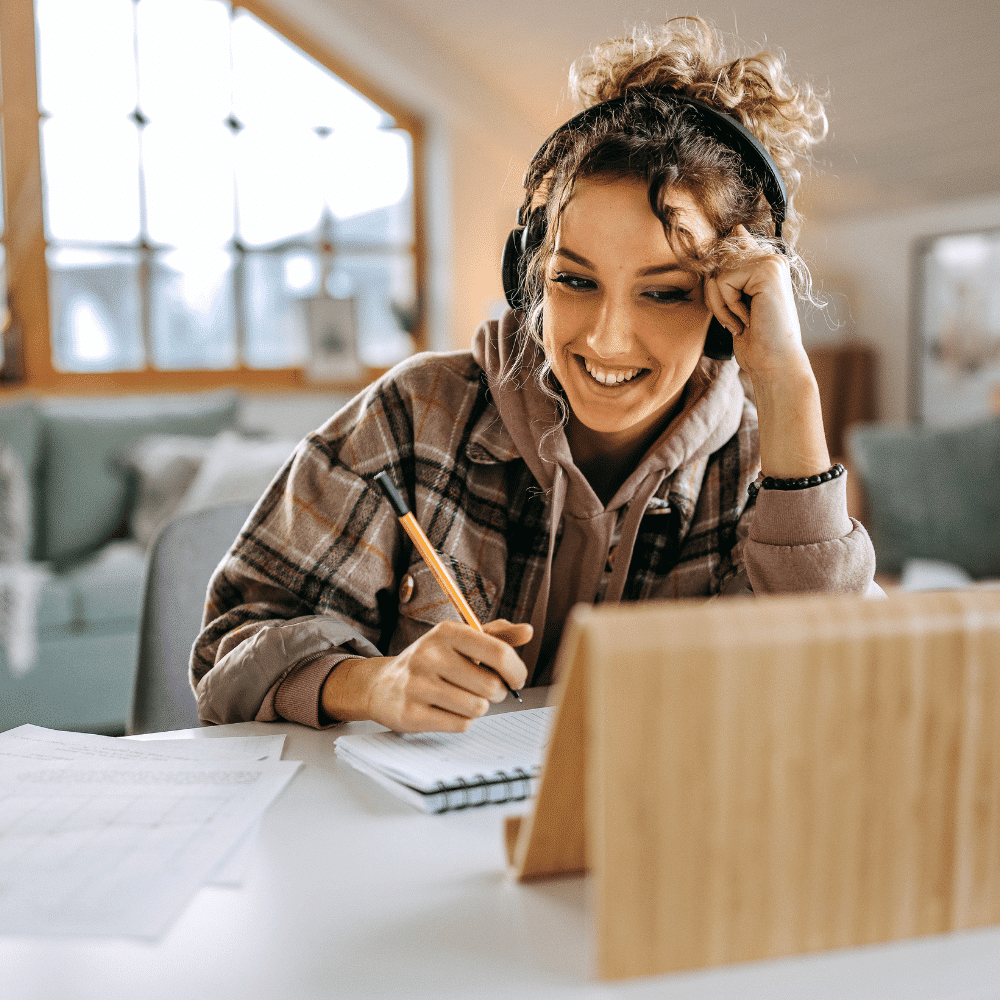 A young woman wearing headphones smiles while taking notes in a notebook, sitting at a desk with a tablet propped up in front of her in a cozy, sunlit room.