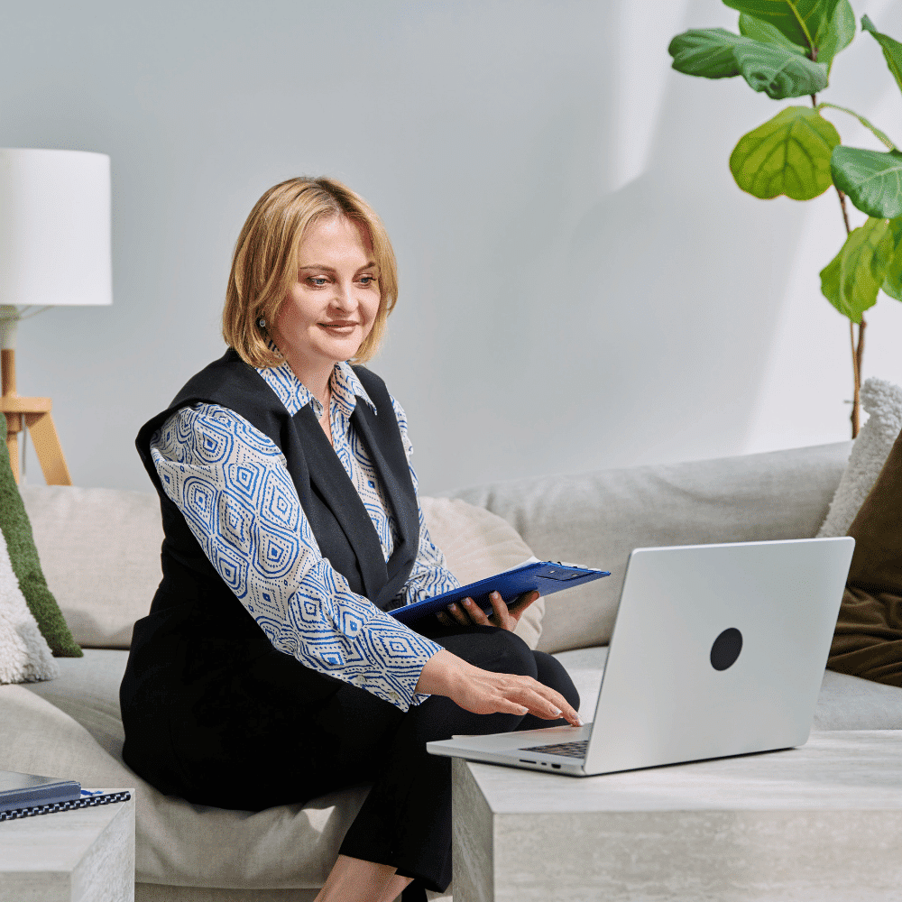 A woman sits on a light-colored sofa, using a laptop on a coffee table. She is holding a blue folder and smiling. There are green plants and a lamp in the background, creating a bright, modern setting.