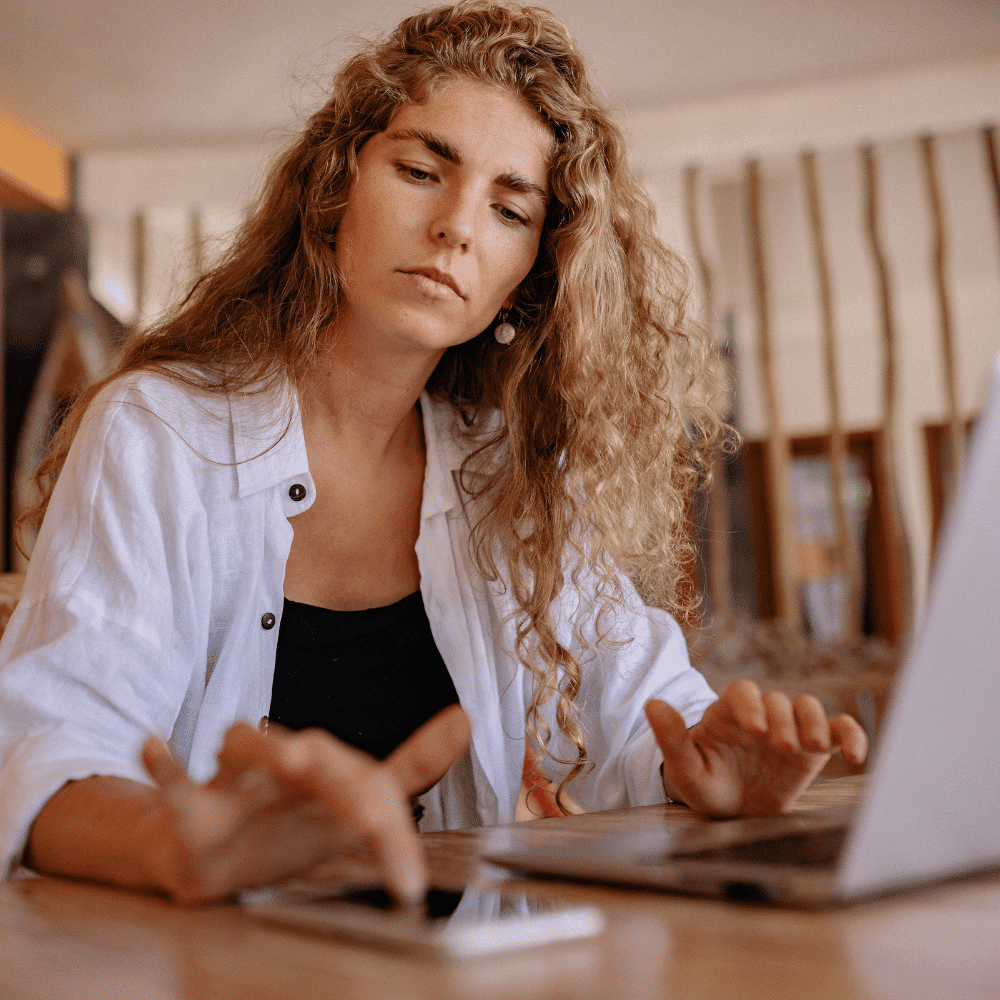 A woman with long curly hair sits at a table, looking at a laptop screen while using a smartphone with her other hand. She appears focused and is wearing a white shirt over a black top.