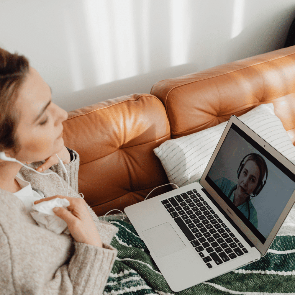 A person sits on a couch with a blanket, holding tissues and video chatting with another person wearing a headset, visible on the laptop screen.