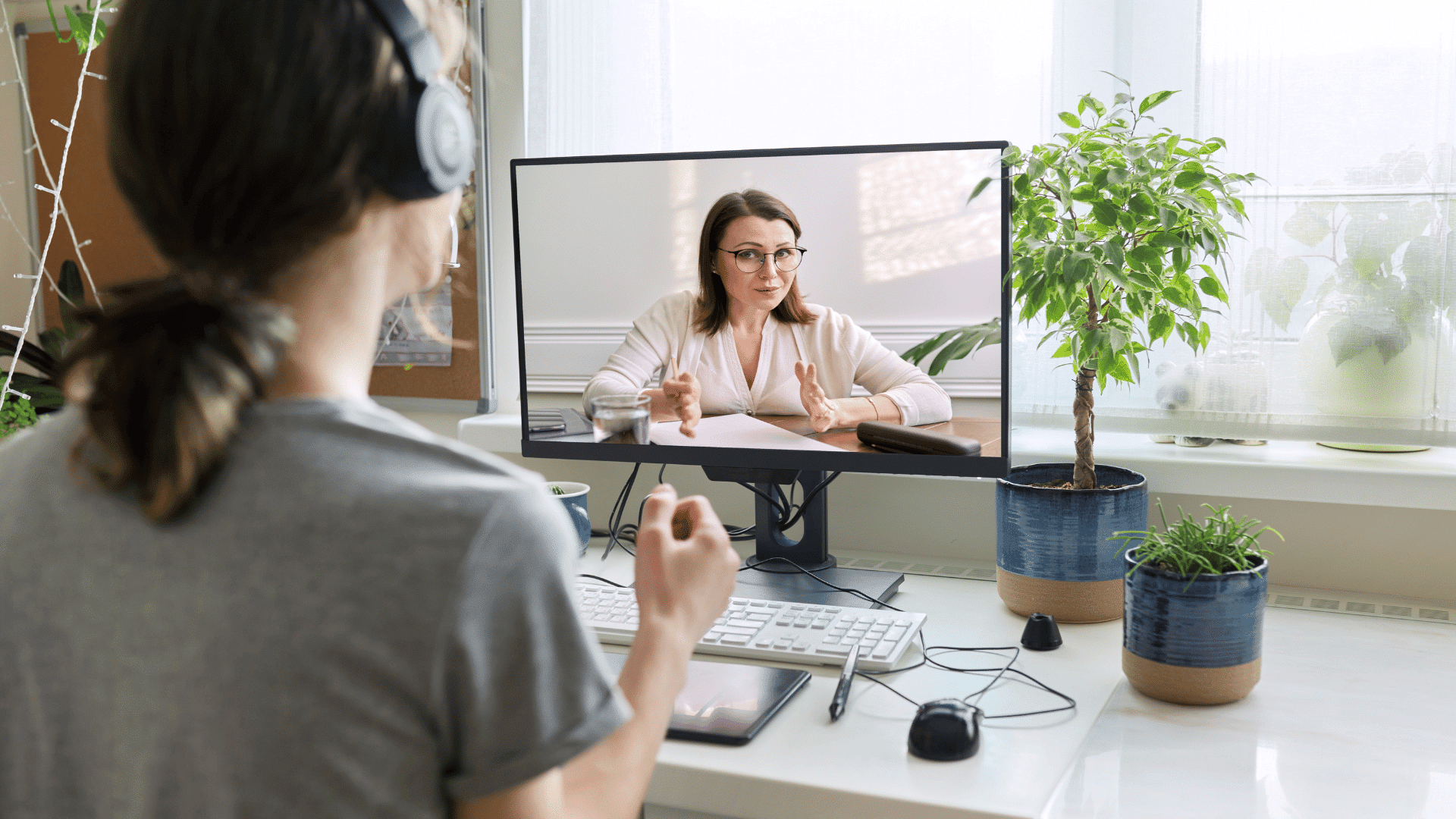 A person wearing headphones sits at a desk, participating in a video call with a woman on a computer screen. The desk is organized and surrounded by potted plants, and natural light streams in through a window.