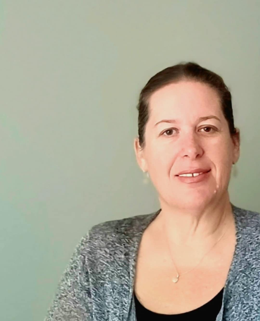 A woman with brown hair pulled back smiles softly at the camera. She is wearing a gray cardigan over a black top, with a plain light green background behind her.