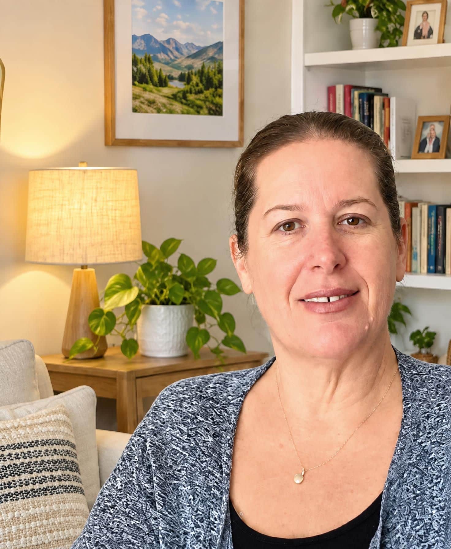 Woman smiling indoors, sitting on a couch with bookshelves, a lamp, and a plant in the background. A framed landscape photo hangs on the wall. The room feels cozy and well-lit.
